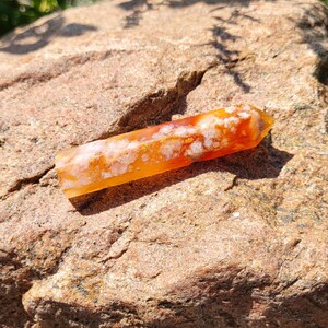 May include: A polished, rectangular crystal with a pointed end. The crystal is a blend of orange, yellow, and white hues, with a translucent quality. It rests on a rough, brown rock surface, illuminated by sunlight.