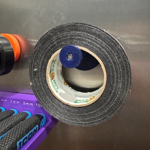 May include: A roll of black Duck Tape with a blue center piece. The tape is mounted on a metal surface. The brand name "DUCK TAPE" is visible on the tape roll. A purple tool holder with "ICON" is in the lower left.