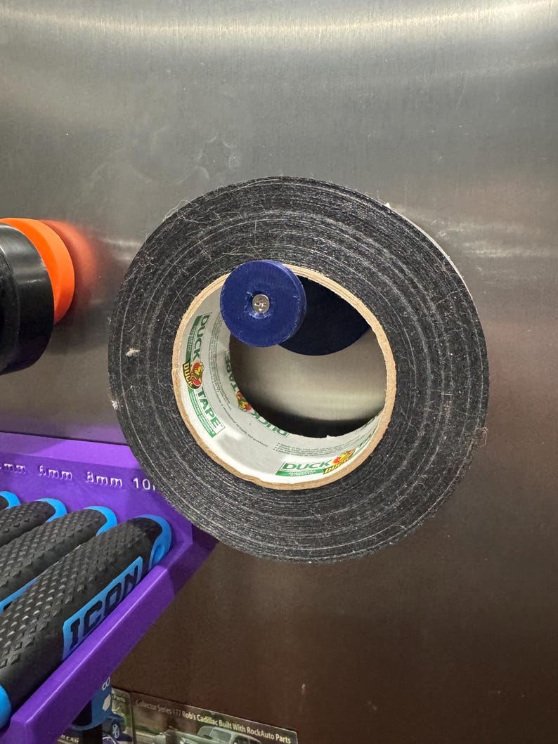 May include: A roll of black Duck Tape with a blue center piece. The tape is mounted on a metal surface. The brand name "DUCK TAPE" is visible on the tape roll. A purple tool holder with "ICON" is in the lower left.