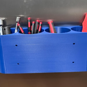 Könnte beinhalten: Ein blauer, 3D-gedruckter Werkzeugorganizer mit mehreren Fächern. Er enthält verschiedene Werkzeuge, darunter Schraubendreher mit roten Griffen, Steckschlüssel und einen roten Schaber. Der Organizer hat ein gebogenes Design und ist auf einer Oberfläche montiert.