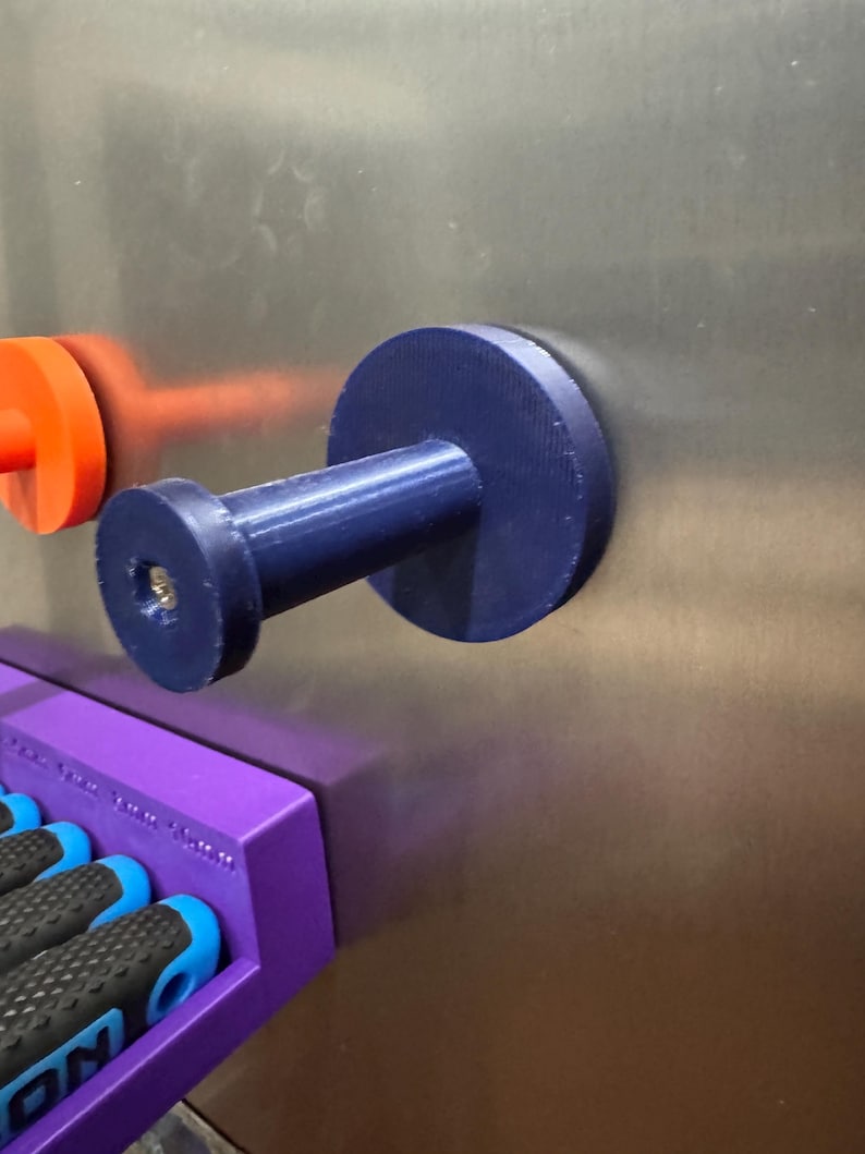 May include: A dark blue magnetic hook with a cylindrical stem and a circular base is attached to a metal surface. An orange magnetic disc is visible to the left. A purple tool holder is below the hook, with the handles of tools visible.