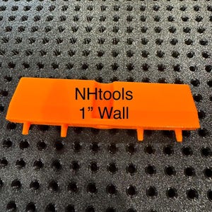 Puede incluir: Herramienta de color naranja brillante con el texto "NHtools 1" Wall". La herramienta tiene forma rectangular con cuatro pequeñas protuberancias en la parte inferior. La herramienta está sobre un fondo negro texturizado.