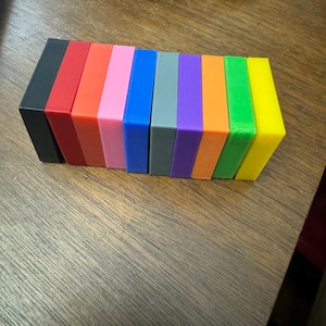 Puede incluir: Una fila de diez bloques rectangulares peque&ntilde;os en varios colores, incluyendo negro, rojo, naranja, rosa, azul, gris, morado, naranja, verde y amarillo.