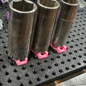 Puede incluir: Tres llaves de vaso plateadas con mangos de goma negra est&aacute;n organizadas en una placa perforada negra con soportes rosas y morados.