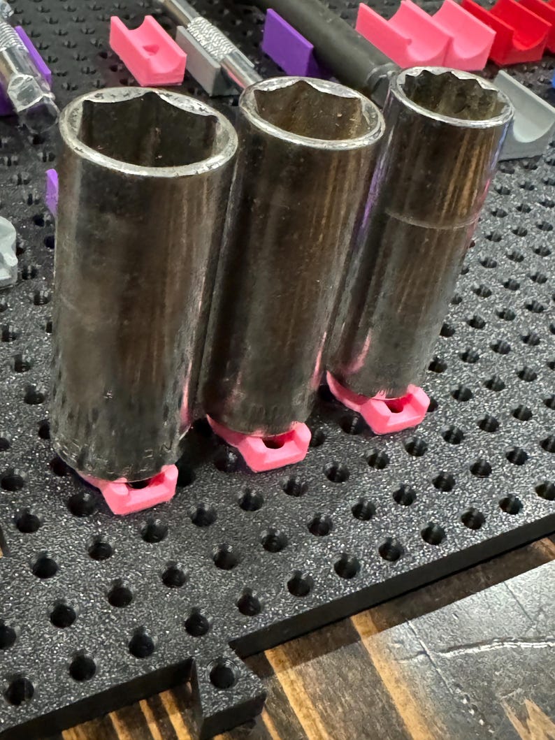 Puede incluir: Tres llaves de vaso plateadas con mangos de goma negra est&aacute;n organizadas en una placa perforada negra con soportes rosas y morados.