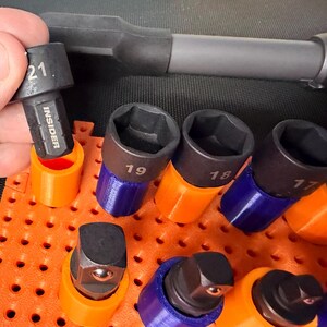 Puede incluir: Un juego de vasos de impacto de metal negro etiquetados con los tamaños 17, 18, 19 y 21, incluyendo uno marcado como INSIDER. Los vasos se sujetan en soportes de plástico naranja y azul sobre una base de tablero perforado naranja.