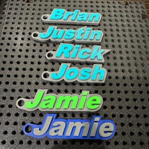 May include: Six personalized keychains with names "Brian", "Justin", "Rick", "Josh", "Jamie", and "Jamie" printed in different colors on a gray background.