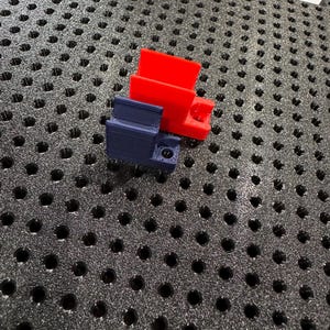 May include: Two 3D-printed plastic components, one red and one dark blue, are displayed on a textured black surface. The red piece has a rectangular shape with a protruding section. The blue piece has a more complex design with a small circular opening.