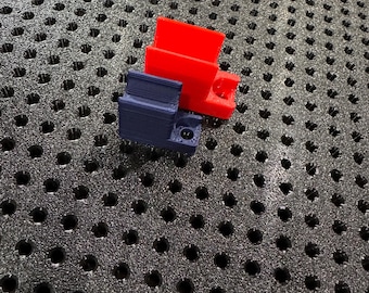 Puede incluir: Dos componentes de plástico impresos en 3D, uno rojo y otro azul oscuro, se muestran sobre una superficie negra texturizada. La pieza roja tiene forma rectangular con una sección saliente. La pieza azul tiene un diseño complejo con una pequeña abertura circular.
