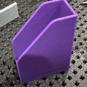 Puede incluir: Un contenedor en ángulo morado vibrante impreso en 3D. El diseño de tapa abierta presenta una base rectangular y lados en ángulo, creando un espacio de almacenamiento. El contenedor está hecho de un material sólido y liso. El artículo está sobre una superficie texturizada.