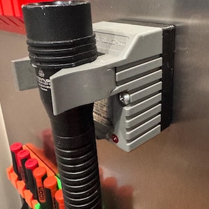 Könnte beinhalten: Eine schwarze Streamlight LED-Taschenlampe in einer grauen Kunststoff-Ladestation, die an einer Metalloberfläche befestigt ist. Die Taschenlampe hat einen strukturierten Griff und ein Warnsymbol für heiße Oberflächen. Eine rote Leuchte ist sichtbar.