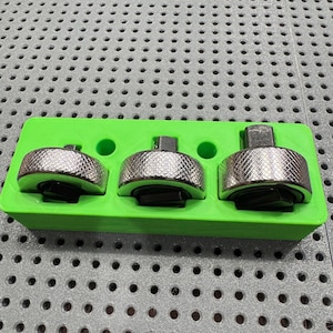 Op de afbeelding: Drie zilveren ratelkoppen met getextureerde grepen zijn in een felgroene houder geplaatst. De gereedschappen zijn ontworpen voor mechanisch werk. De houder heeft drie ronde gaten en een rechthoekige vorm. De achtergrond is een grijs, geperforeerd oppervlak.