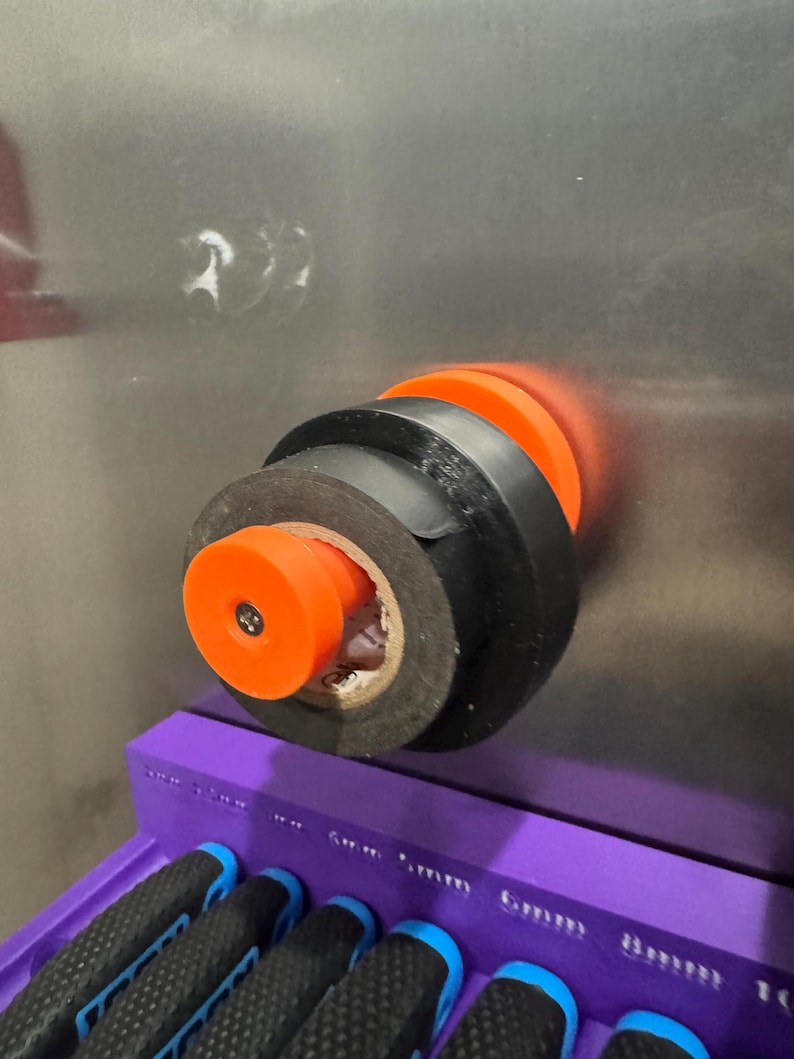 May include: An orange and black tool holder with black electrical tape wrapped around it. The tool holder is mounted on a metallic surface. A purple tool organizer with several tools is visible in the foreground.
