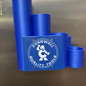 Puede incluir: Un organizador de herramientas azul con múltiples compartimentos cilíndricos de diferentes alturas. El organizador presenta una base rectangular con el logotipo "Cornwell Quality Tools" en blanco. El logotipo es un círculo con una figura sosteniendo una llave.