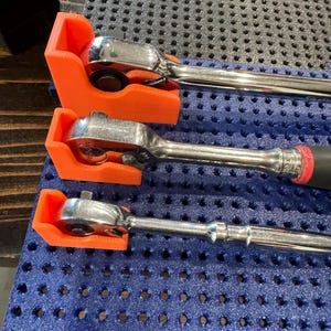May include: Three chrome ratchets are held in orange 3D-printed holders. The ratchets are on a blue perforated surface. The tools are designed for mechanical work and are commonly used in automotive repair and construction.