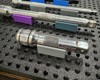 Puede incluir: Primer plano de un tablero de clavijas con cuatro llaves de vaso de metal plateado colgadas. Las llaves se mantienen en su lugar mediante clips de plástico de colores. El tablero de clavijas es negro y tiene un patrón de cuadrícula.