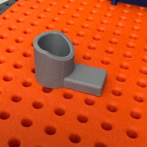 Könnte beinhalten: Ein grauer Plastikhalter mit einer runden Öffnung oben und einer flachen Basis. Der Halter steht auf einer orangefarbenen Oberfläche mit einem Gittermuster.