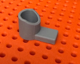 Puede incluir: Un soporte de plástico gris con una abertura redonda en la parte superior y una base plana. El soporte está sobre una superficie naranja con un patrón de cuadrícula.