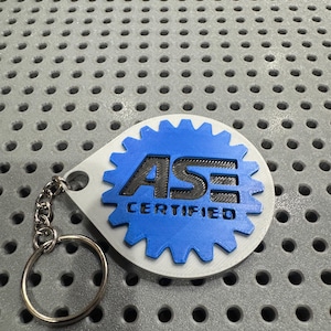 May include: A white and blue keychain with a gear-shaped charm. The charm has the text "ASE CERTIFIED" in black lettering.