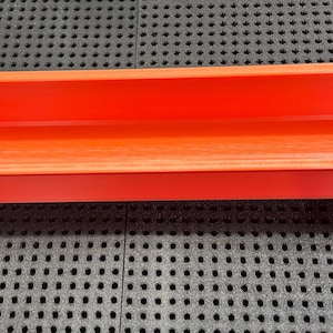Könnte beinhalten: Ein leuchtend orangefarbener rechteckiger Pflanzkübel mit abgerundeten Ecken. Der Pflanzkübel hat eine glatte Innenseite und zwei kleine Abzugslöcher. Er steht auf einer schwarzen Oberfläche mit einem Gittermuster. Ca. 25 cm lang.