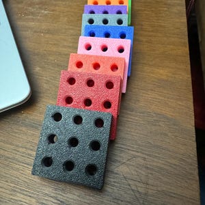 Puede incluir: Un conjunto de diez bloques cuadrados coloridos con agujeros, apilados en forma de pir&aacute;mide. Los bloques est&aacute;n hechos de un material texturizado y son de color rojo, naranja, amarillo, verde, azul, morado, rosa, rosa claro, gris y negro.