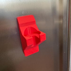 Op de afbeelding: Een felrode, 3D-geprinte houder, waarschijnlijk voor een beker of fles, bevestigd aan een roestvrijstalen oppervlak. De houder heeft een gebogen ontwerp met een cirkelvormige opening en een rechthoekige basis. Het object is gemaakt van een solide, ondoorzichtig materiaal.