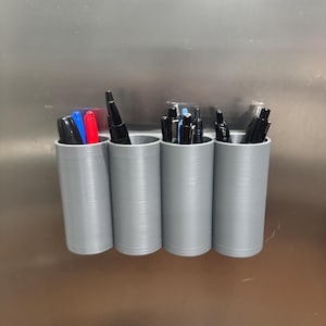 Könnte beinhalten: Ein grauer, 3D-gedruckter Stifthalter mit vier zylindrischen Fächern. Jedes Fach ist mit Stiften und Markern in verschiedenen Farben gefüllt, darunter Schwarz, Blau und Rot. Der Halter ist auf einer metallischen Oberfläche montiert.
