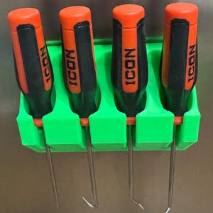 Op de afbeelding: Vier haak- en pickgereedschappen met oranje en zwarte handvatten, met het label "ICON", worden in een groene plastic organizer gehouden. De gereedschappen hebben gebogen metalen punten. De organizer is op een metalen oppervlak gemonteerd.