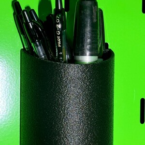 May include: A black pen holder with a textured surface holds a variety of black pens, including a Pilot G-2 07 pen.