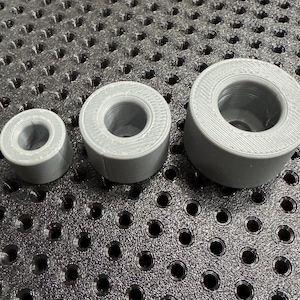 Op de afbeelding: Drie grijze, cilindrische objecten met een centraal gat, gerangschikt van klein naar groot. De objecten hebben een geribbelde textuur aan de buitenrand en lijken 3D-geprint te zijn. De objecten staan op een zwart, getextureerd oppervlak.