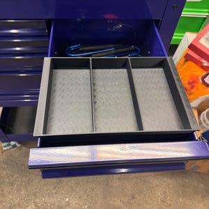 May include: A black plastic drawer organizer with three rectangular compartments featuring a textured diamond plate pattern on the base, positioned inside an open purple metal tool chest drawer with a smooth, glossy finish and a locking mechanism.