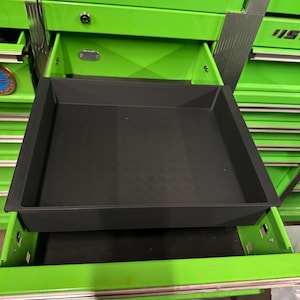 May include: A black, square, shallow plastic tray with a smooth interior base and raised edges, positioned inside an open, bright green metal tool chest drawer. The tray is designed to fit within the drawer compartment for organized storage.