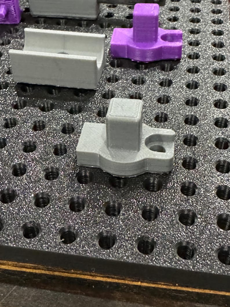 Puede incluir: Dos piezas de pl&aacute;stico impresas en 3D, una gris y otra morada, est&aacute;n en un tablero de clavijas negro. La pieza gris tiene una parte superior cuadrada y una base redonda. La pieza morada tiene una parte superior cuadrada y una base redonda con un agujero en el centro.