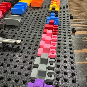 Puede incluir: Primer plano de un tablero de clavijas con varias piezas de pl&aacute;stico de colores dispuestas en filas. Las piezas son de diferentes formas y tama&ntilde;os, y todas est&aacute;n sujetas al tablero de clavijas con peque&ntilde;as clavijas. El tablero de clavijas es negro y tiene un patr&oacute;n de cuadr&iacute;cula.