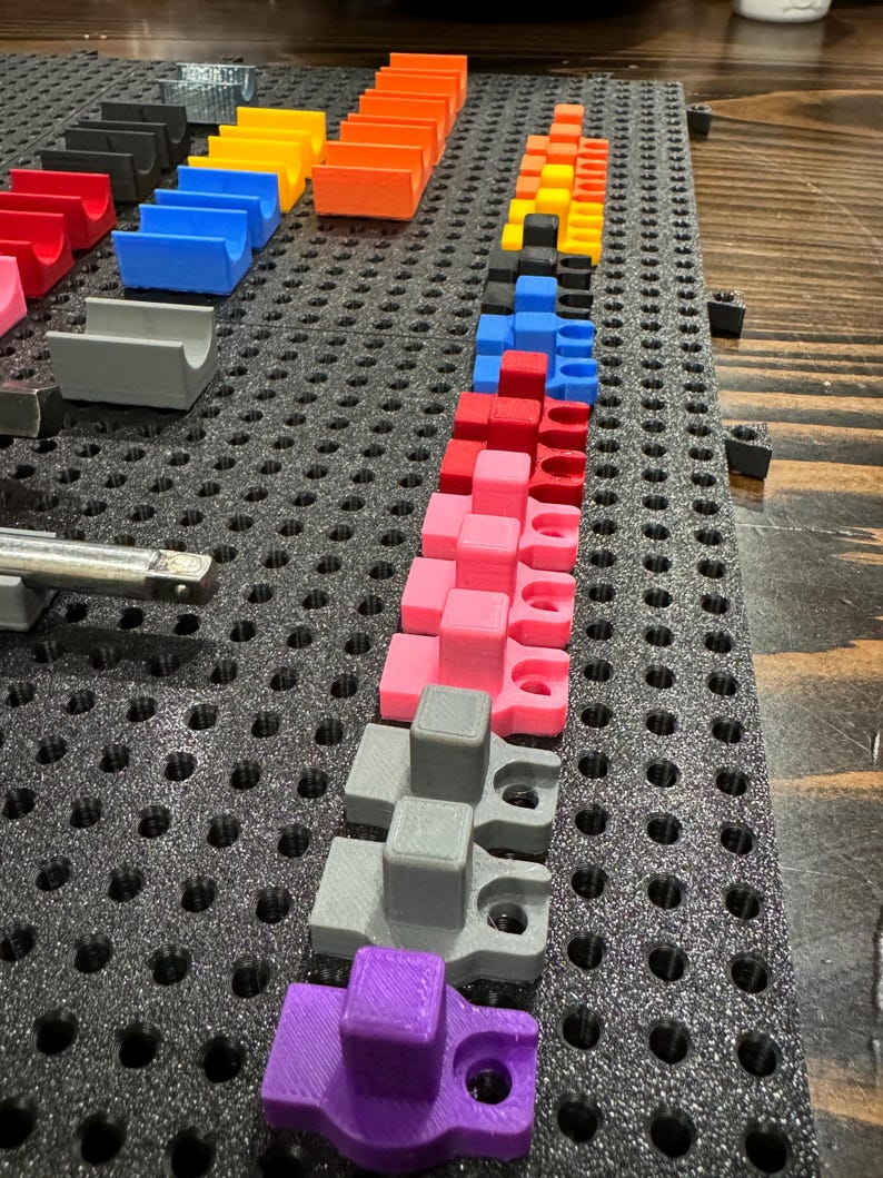 Puede incluir: Primer plano de un tablero de clavijas con varias piezas de pl&aacute;stico de colores dispuestas en filas. Las piezas son de diferentes formas y tama&ntilde;os, y todas est&aacute;n sujetas al tablero de clavijas con peque&ntilde;as clavijas. El tablero de clavijas es negro y tiene un patr&oacute;n de cuadr&iacute;cula.