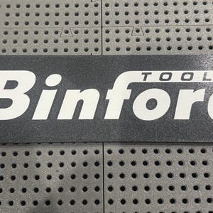 Puede incluir: Una alfombrilla rectangular gris oscuro con esquinas redondeadas. El texto blanco "Binford TOOLS" se muestra de forma destacada. La alfombrilla se utiliza probablemente en un taller o garaje.