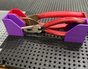Puede incluir: Unos alicates con mango rojo descansan en un soporte para herramientas impreso en 3D de color morado y rojo. Los alicates son de metal y muestran signos de uso. El soporte está sobre una superficie negra texturizada.