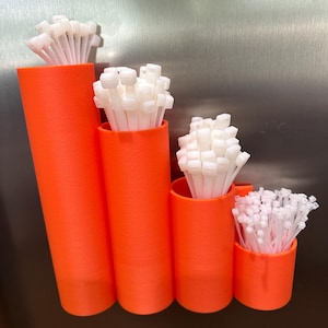 Könnte beinhalten: Vier orangefarbene zylindrische Behälter unterschiedlicher Höhe, jeder gefüllt mit weißen Kabelbindern. Die Behälter sind in einer Reihe angeordnet, der höchste links und der kürzeste rechts. Der Hintergrund ist eine metallische Oberfläche.