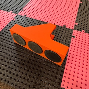 Könnte beinhalten: Ein orangefarbenes, geometrisches Objekt mit drei schwarzen, kreisförmigen Elementen. Es liegt auf einer Oberfläche aus ineinandergreifenden schwarzen und rosa Schaumstofffliesen. Die Fliesen haben ein strukturiertes, perforiertes Design.