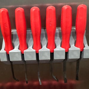 Op de afbeelding: Een set van zes koevoeten met rode handgrepen in een grijze houder. De handgrepen zijn gemarkeerd met "USA". De koevoeten hebben een gebogen of plat lemmet. Deze gereedschappen zijn ontworpen om objecten te wrikken, op te tillen en te verwijderen.