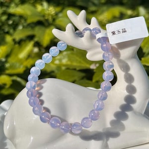 May include: A beaded bracelet with light blue and lavender-colored beads. The bracelet is displayed on a white ceramic deer figurine. A white tag with Chinese text is attached to the figurine.