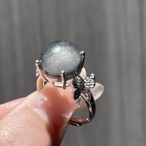Puede incluir: Un anillo plateado con una piedra preciosa gris redonda y grande con un efecto brillante. El anillo tiene un dise&ntilde;o de mariposa en la banda. La piedra preciosa est&aacute; sujeta por cuatro garras.