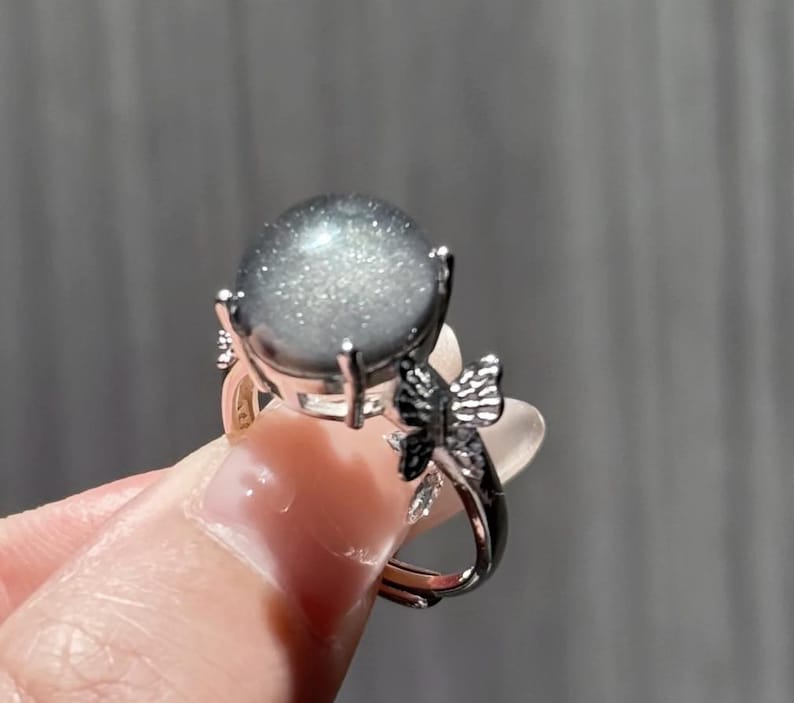 Puede incluir: Un anillo plateado con una piedra preciosa gris redonda y grande con un efecto brillante. El anillo tiene un dise&ntilde;o de mariposa en la banda. La piedra preciosa est&aacute; sujeta por cuatro garras.