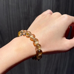 K&ouml;nnte beinhalten: Ein Armband aus goldbraunen Perlen mit einer Mischung aus klaren und undurchsichtigen Steinen. Die runden, polierten Perlen werden am Handgelenk getragen. Die Perlen schimmern leicht und variieren von hellbernstein bis dunkelbraun.
