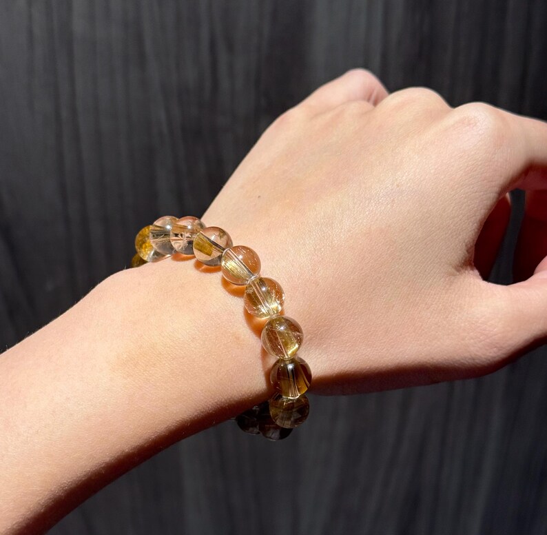K&ouml;nnte beinhalten: Ein Armband aus goldbraunen Perlen mit einer Mischung aus klaren und undurchsichtigen Steinen. Die runden, polierten Perlen werden am Handgelenk getragen. Die Perlen schimmern leicht und variieren von hellbernstein bis dunkelbraun.
