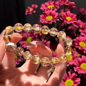 K&ouml;nnte beinhalten: Ein Armband aus klaren Quarzperlen mit goldenen Einschl&uuml;ssen. Die runden Perlen sind aufgereiht und erzeugen einen schimmernden Effekt. Das Armband wird vor einem Hintergrund aus rosa Blumen gehalten.