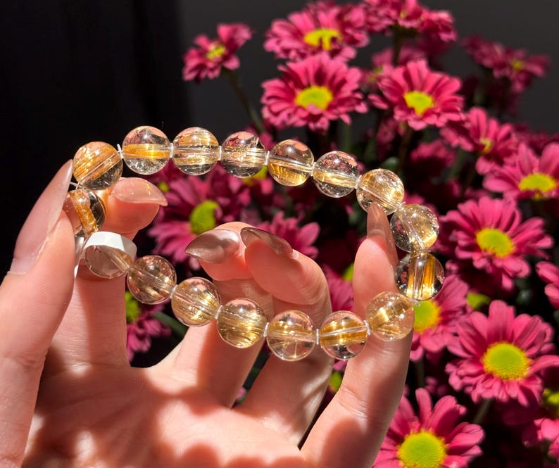 K&ouml;nnte beinhalten: Ein Armband aus klaren Quarzperlen mit goldenen Einschl&uuml;ssen. Die runden Perlen sind aufgereiht und erzeugen einen schimmernden Effekt. Das Armband wird vor einem Hintergrund aus rosa Blumen gehalten.