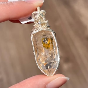 Op de afbeelding: Een heldere kwartskristallen hanger met een zilveren draadwikkeling. Het kristal heeft insluitsels van gele en zwarte mineralen. De hanger wordt tussen twee vingers gehouden.