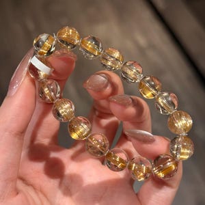 K&ouml;nnte beinhalten: Ein Armband aus klarem Quarz mit goldenen Rutil-Einschl&uuml;ssen. Die runden Perlen haben eine polierte Oberfl&auml;che, die einen funkelnden Effekt erzeugt. Das Armband wird von einer Person gehalten, die die nat&uuml;rliche Sch&ouml;nheit und die einzigartigen Muster in jeder Perle zeigt.