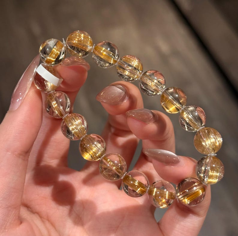 K&ouml;nnte beinhalten: Ein Armband aus klarem Quarz mit goldenen Rutil-Einschl&uuml;ssen. Die runden Perlen haben eine polierte Oberfl&auml;che, die einen funkelnden Effekt erzeugt. Das Armband wird von einer Person gehalten, die die nat&uuml;rliche Sch&ouml;nheit und die einzigartigen Muster in jeder Perle zeigt.
